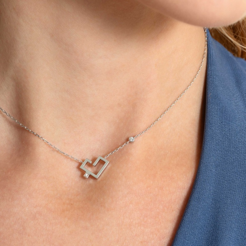 A silver-toned necklace with a heart-shaped pendant and solitaire accent rests against a neck beside a blue garment.