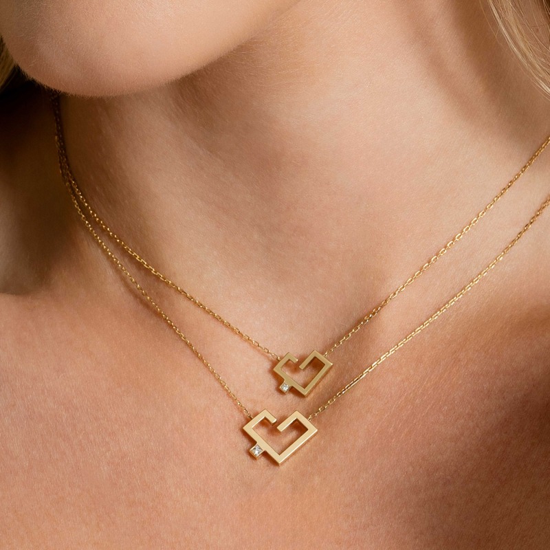 Two layered gold necklaces rest on a neck, each with a heart-shaped Hubb pendant and small sparkling accent.