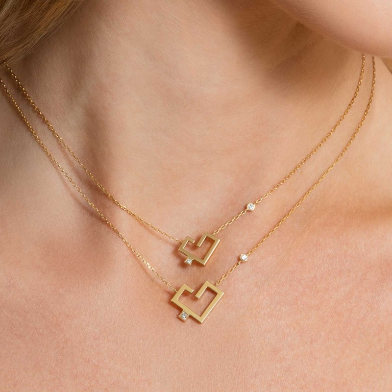 Two layered gold Hubb necklaces on a model’s neck, each with a geometric heart pendant and small solitaire stones.
