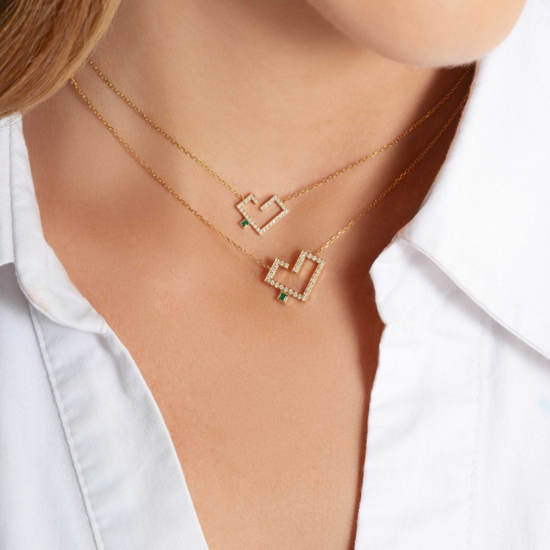 Two delicate gold necklaces with geometric diamond pendants and green accents rest on a woman’s neck above a white shirt.