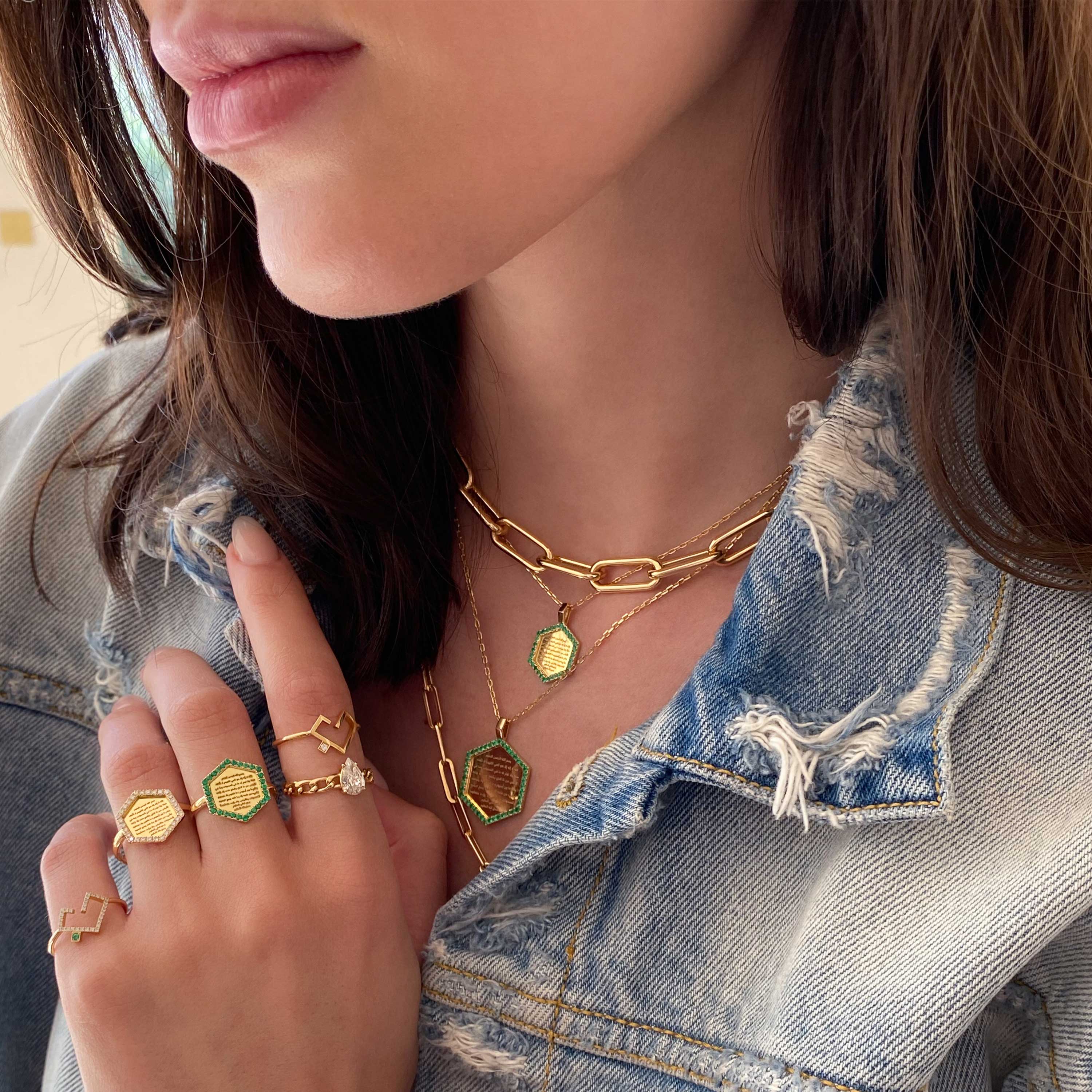 Green emerald Ayat Al-Kursi ring in gold-tone with Quranic inscription, worn with layered chains and denim in a close hand view.
