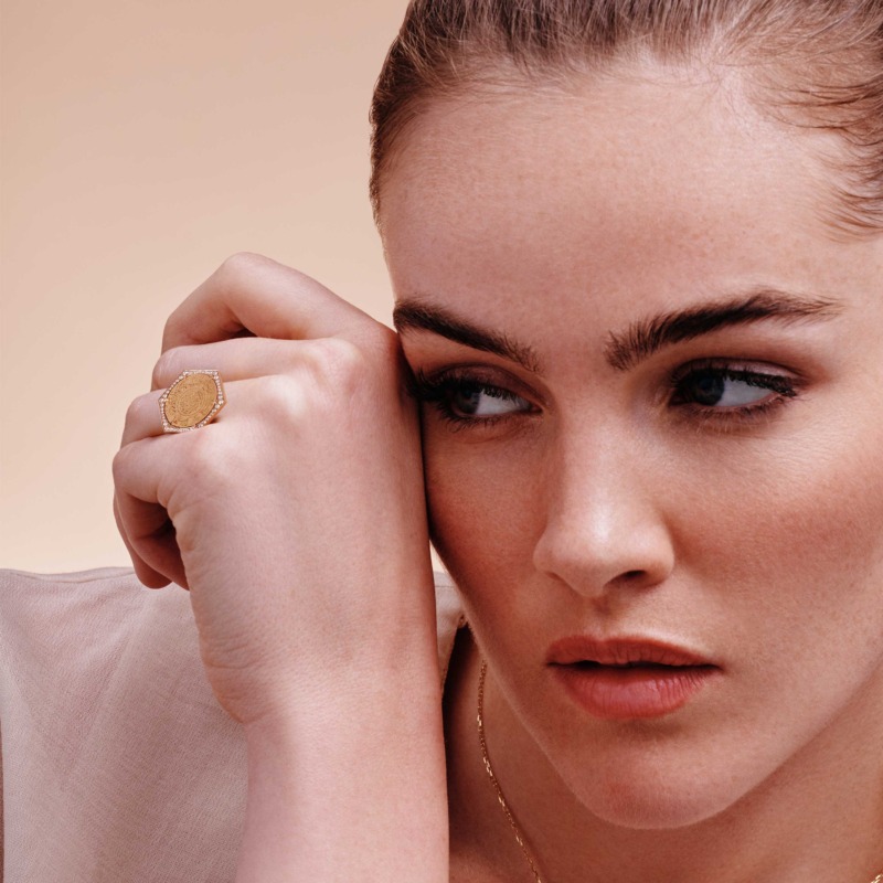 Ginea Ring in diamond frame with a gold-tone setting, oval engraved face and pavé detail, resting near the face in soft light.