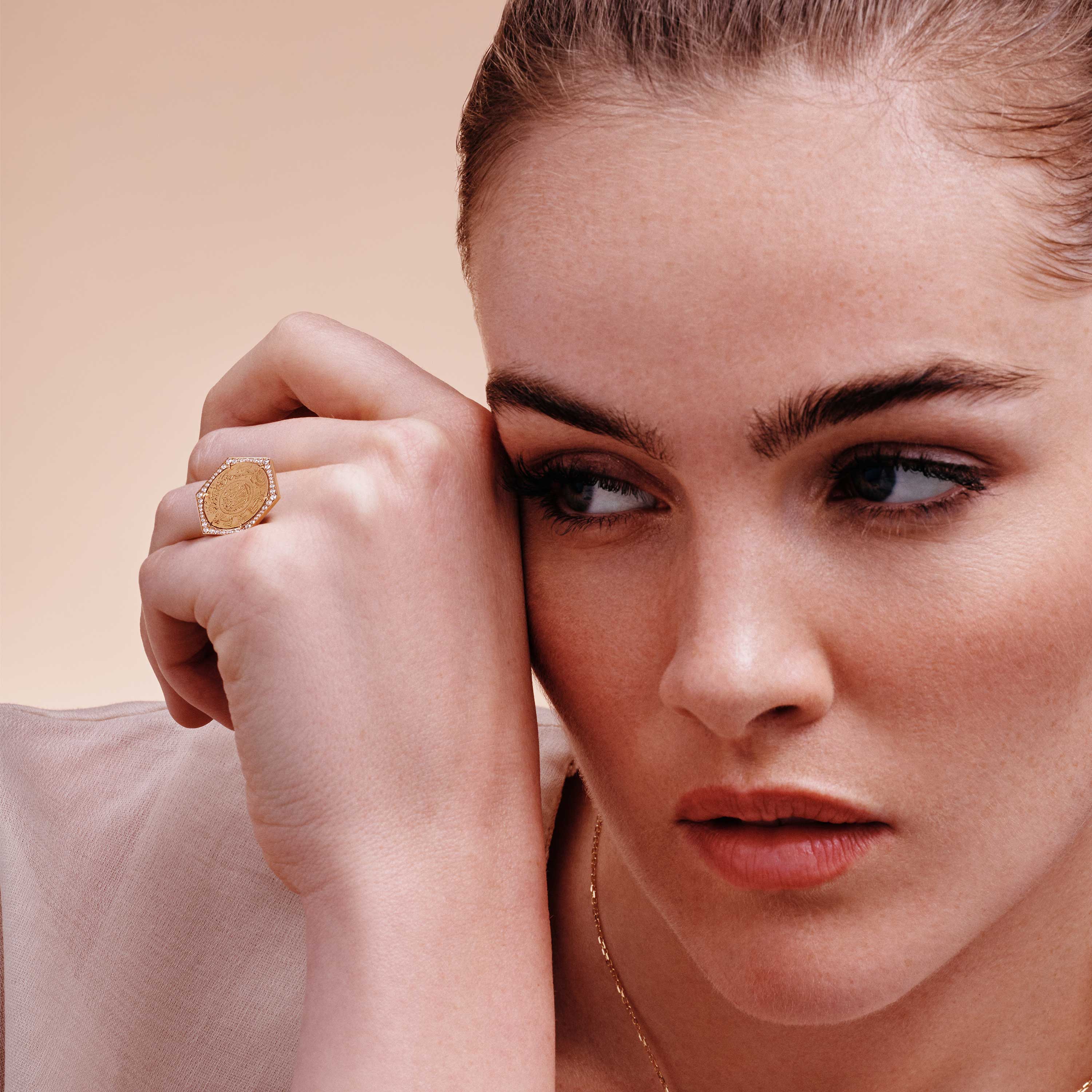Ginea Ring in diamond frame with a gold-tone setting, oval engraved face and pavé detail, resting near the face in soft light.