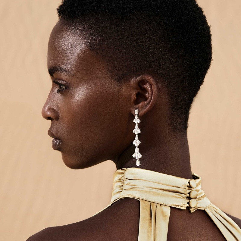 Lotus Evening Earrings by Yataghan Jewellery with dangling lotus charms in silver tones, shown near the ear against a warm beige background.