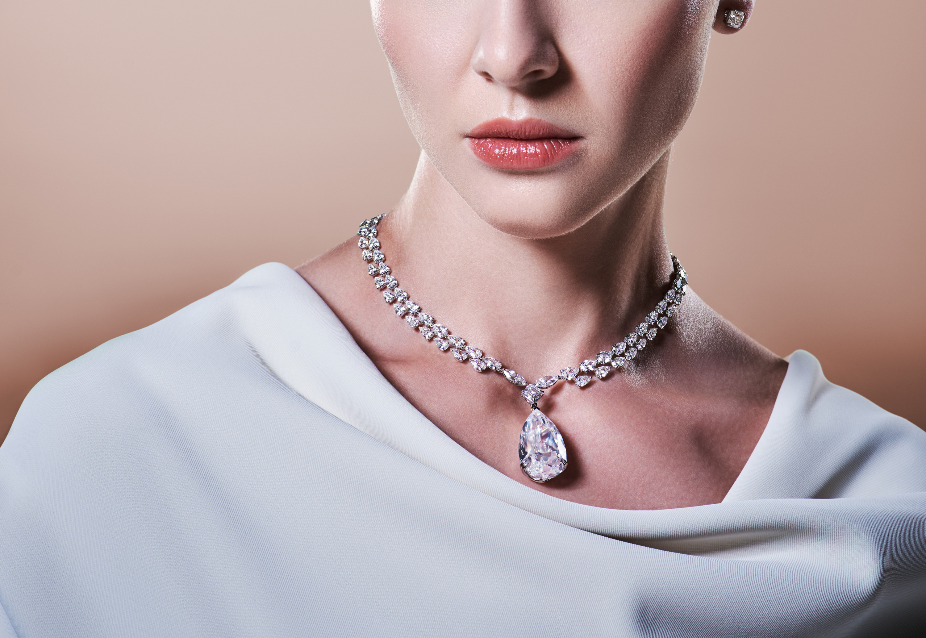 A model wears a diamond necklace with a pear-shaped pendant and matching stud earrings against a soft beige backdrop for Yataghan Jewellery.