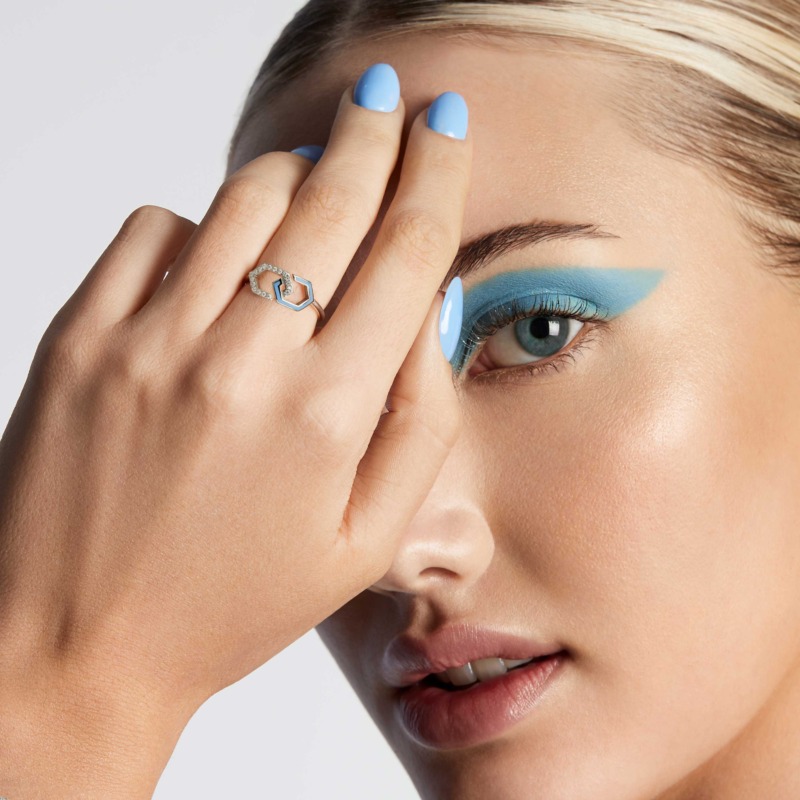 A hand with baby blue nails frames a geometric Infinity ring band, where blue enamel and small diamonds catch the light beside a close-up face.