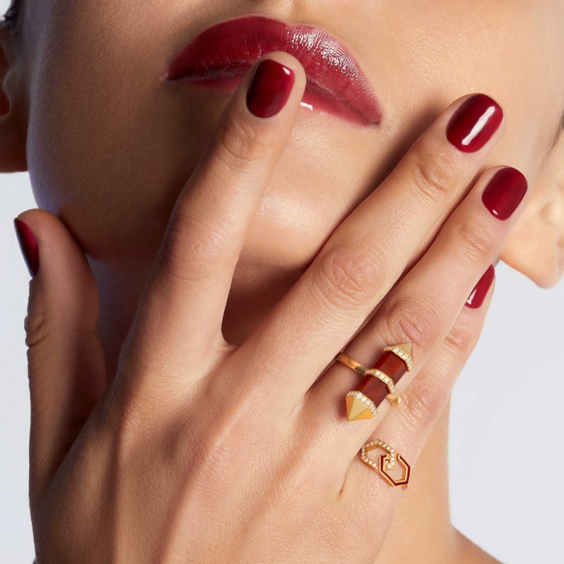 A hand with deep red nails frames a gold Infinity Enamel Diamond Ring Band, whose red enamel and small diamonds add a refined finish.