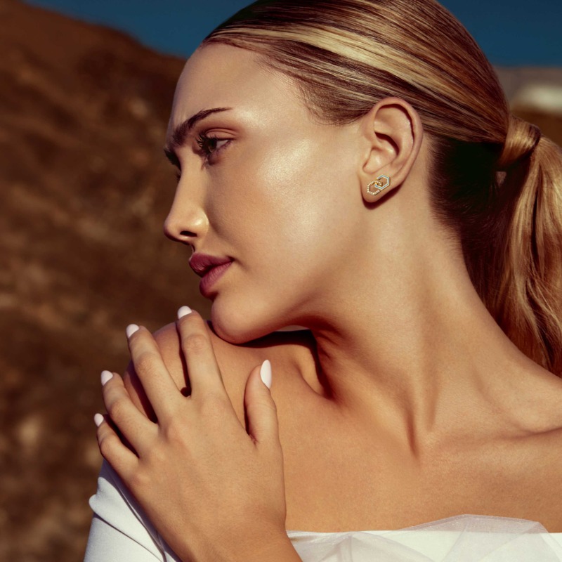 A woman in side profile wears baby blue Infinity enamel diamond stud earrings, with slicked-back hair and a soft outdoor backdrop.