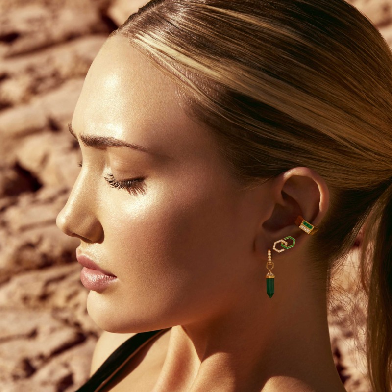 A close profile shows a woman wearing Yataghan Jewellery Infinity Enamel Diamond Stud Earrings with green enamel, gold details, and diamond accents.