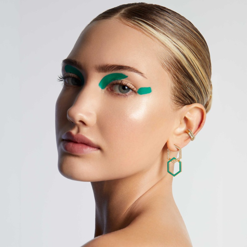A model with slicked-back hair wears turquoise enamel infinity earrings with diamond accents in a close-up portrait against a light background.