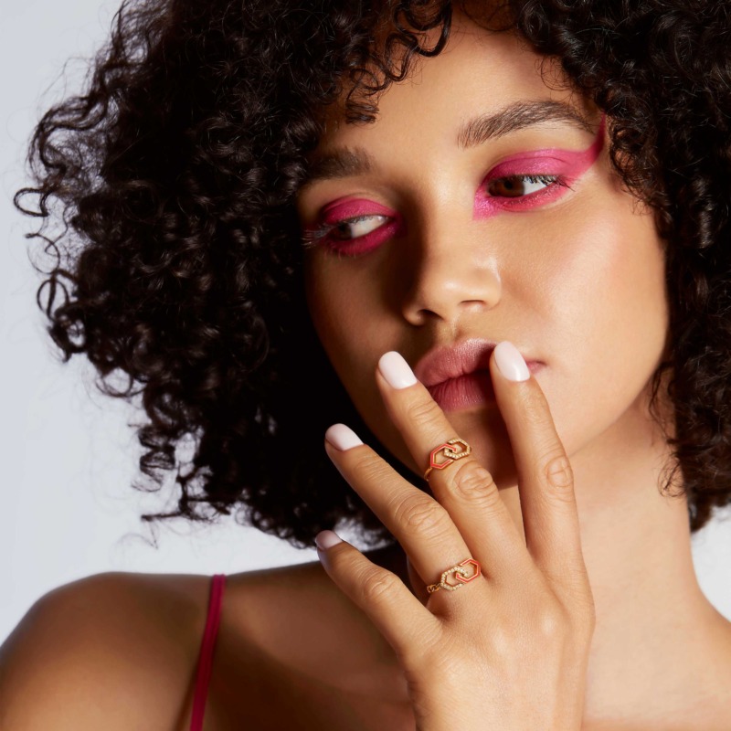 A model with curly hair and bright pink eye makeup holds her hand near her face, showing two gold infinity rings with red enamel and diamonds.