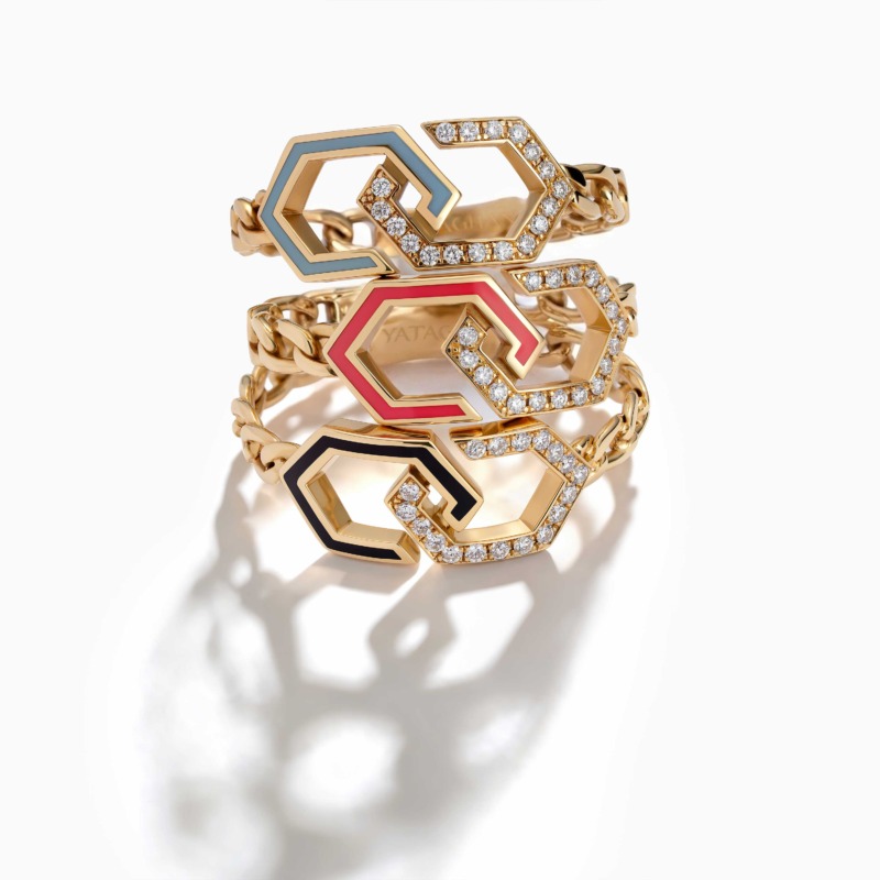 Three stacked gold rings with interlocking geometric links, diamond pavé accents, and blue, red, and black enamel details rest on a white background.