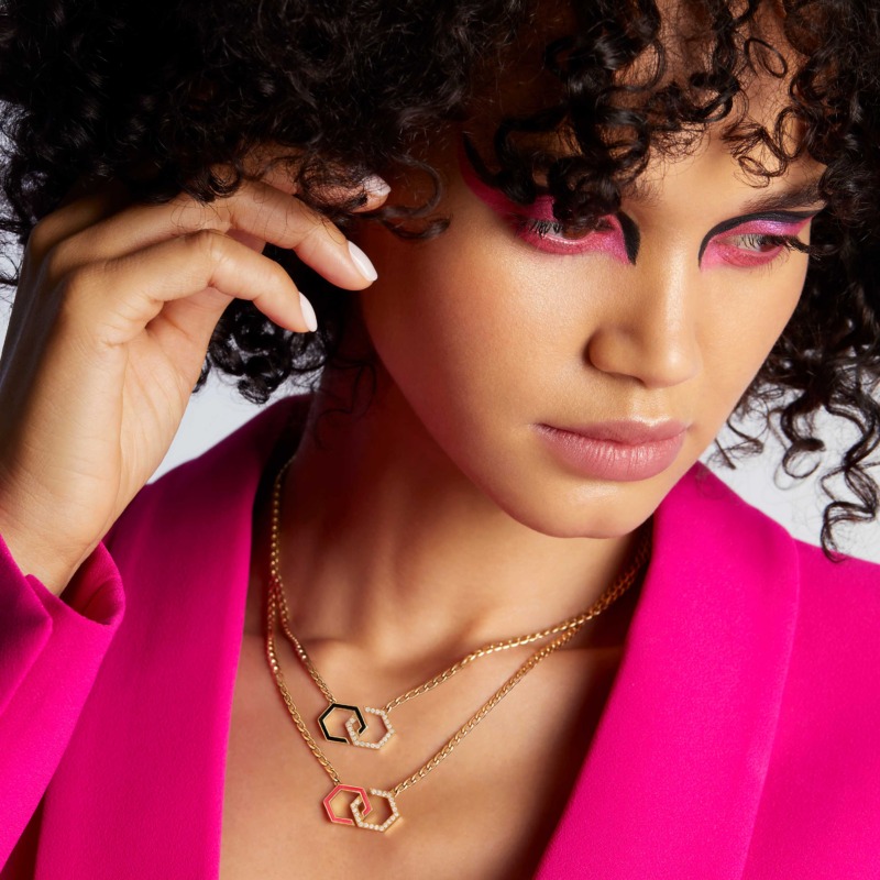 A model with bold pink makeup and a bright fuchsia blazer wears layered gold Infinity enamel necklaces with linked pendant details.