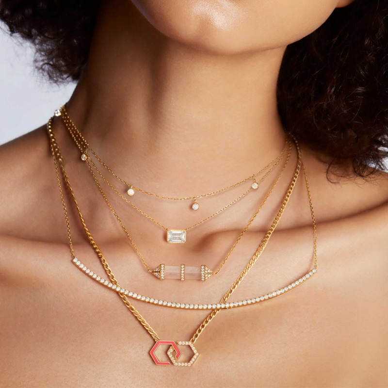 Layered gold chains with diamond accents rest over a model’s neck, centering a pink enamel infinity pendant and geometric charm necklace against soft skin.