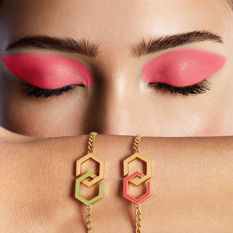 A close-up of an Infinity enamel bracelet in gold with pink and orange accents resting on a wrist beneath bold pink eye makeup.