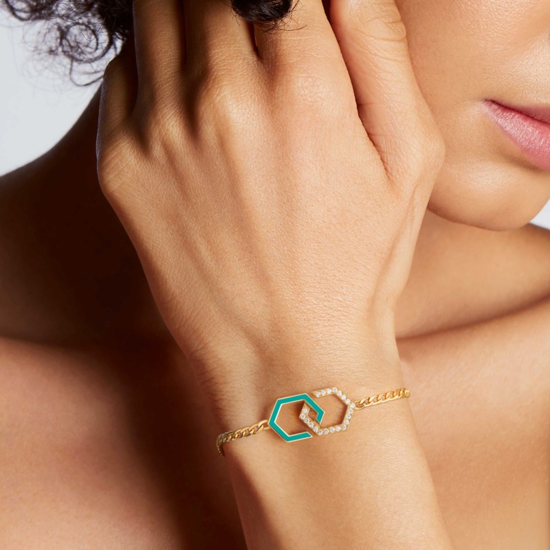 A delicate Infinity bracelet with turquoise enamel and diamond-set links rests on a wrist beside a softly lit face with pink lips and gold chain.