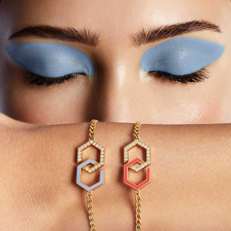 Two gold Infinity enamel bracelets rest across a wrist, each set with pavé diamonds and bright pink or blue enamel in a linked geometric design.