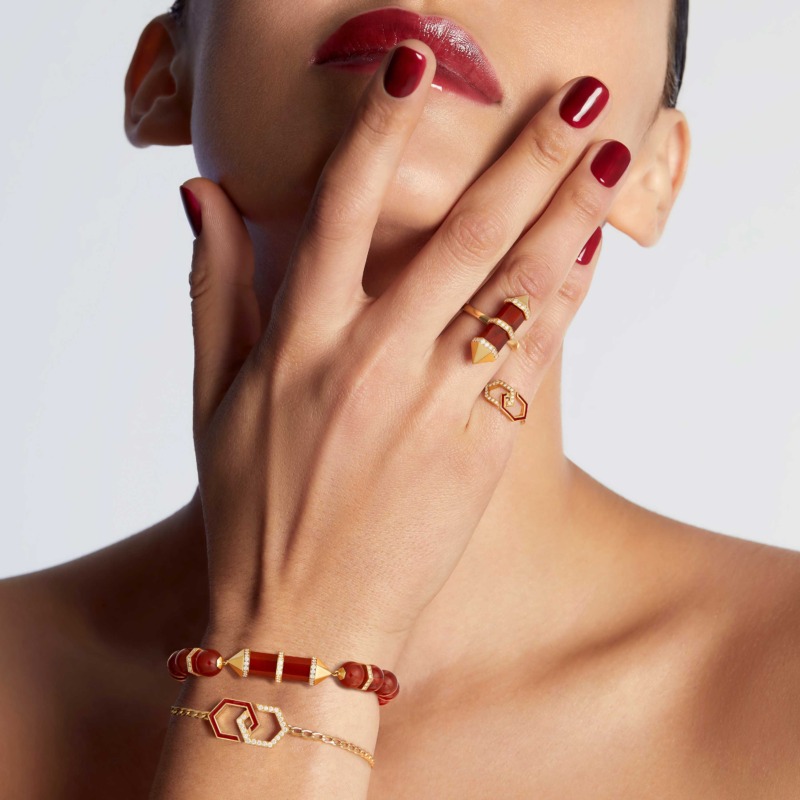 A model with glossy red nails wears an Infinity red enamel bracelet with gold and diamond details across her wrist, beside matching rings and cuffs.