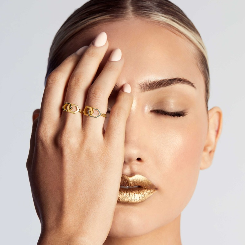 A close-up of a model with metallic gold lips wearing an Infinity enamel unchained gold ring with black enamel links on her hand against a pale background.