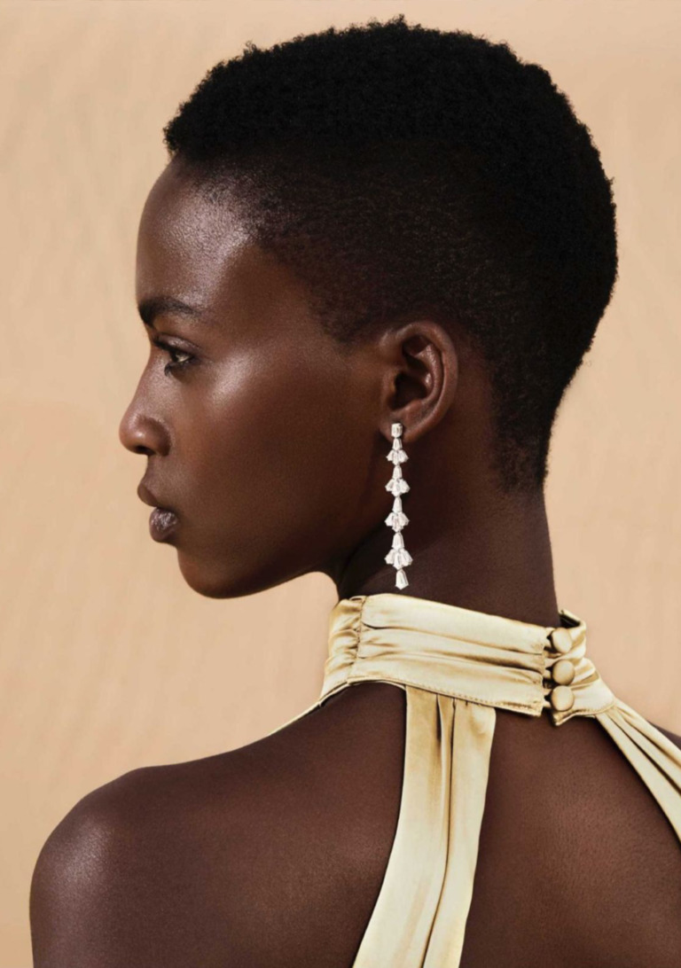 A model in profile wears a long shield-cut diamond dangling earring with a satin halter neckline against a soft beige backdrop.
